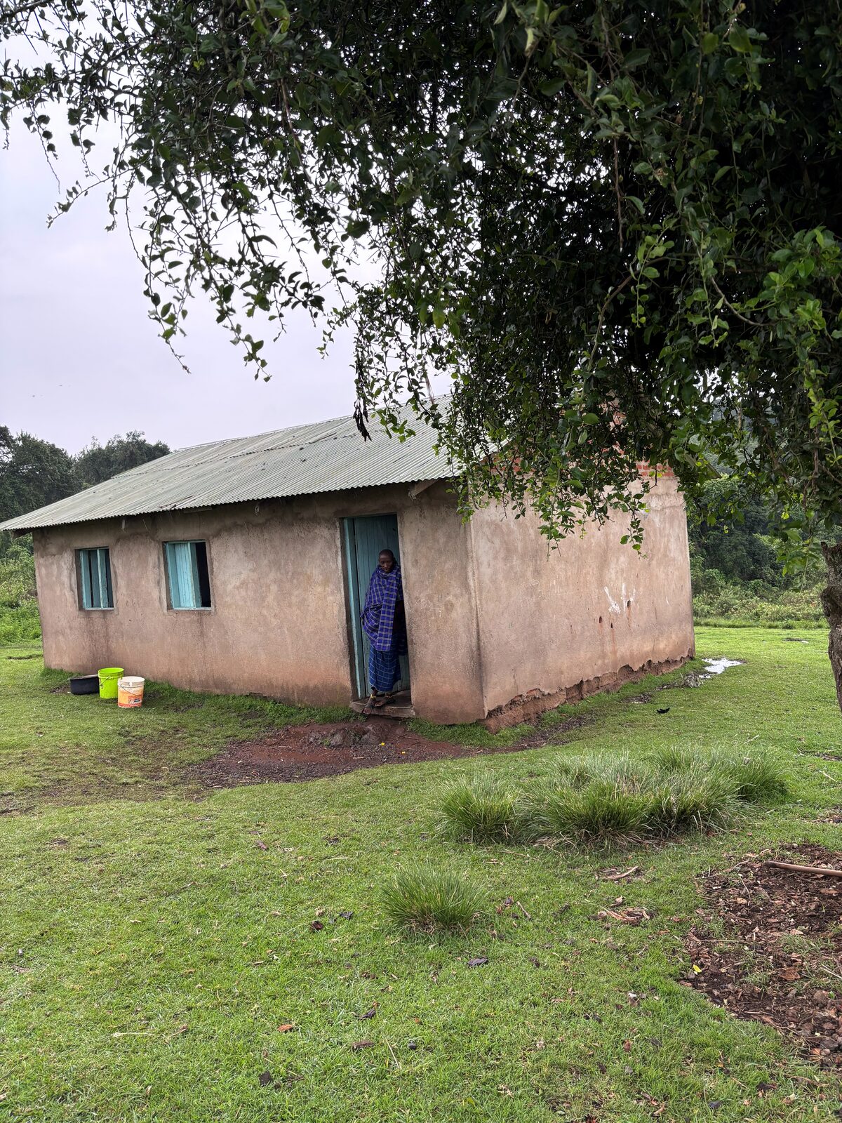 A Boma building under grey skies, a man in traditional Maasai shuka standing in the doorway