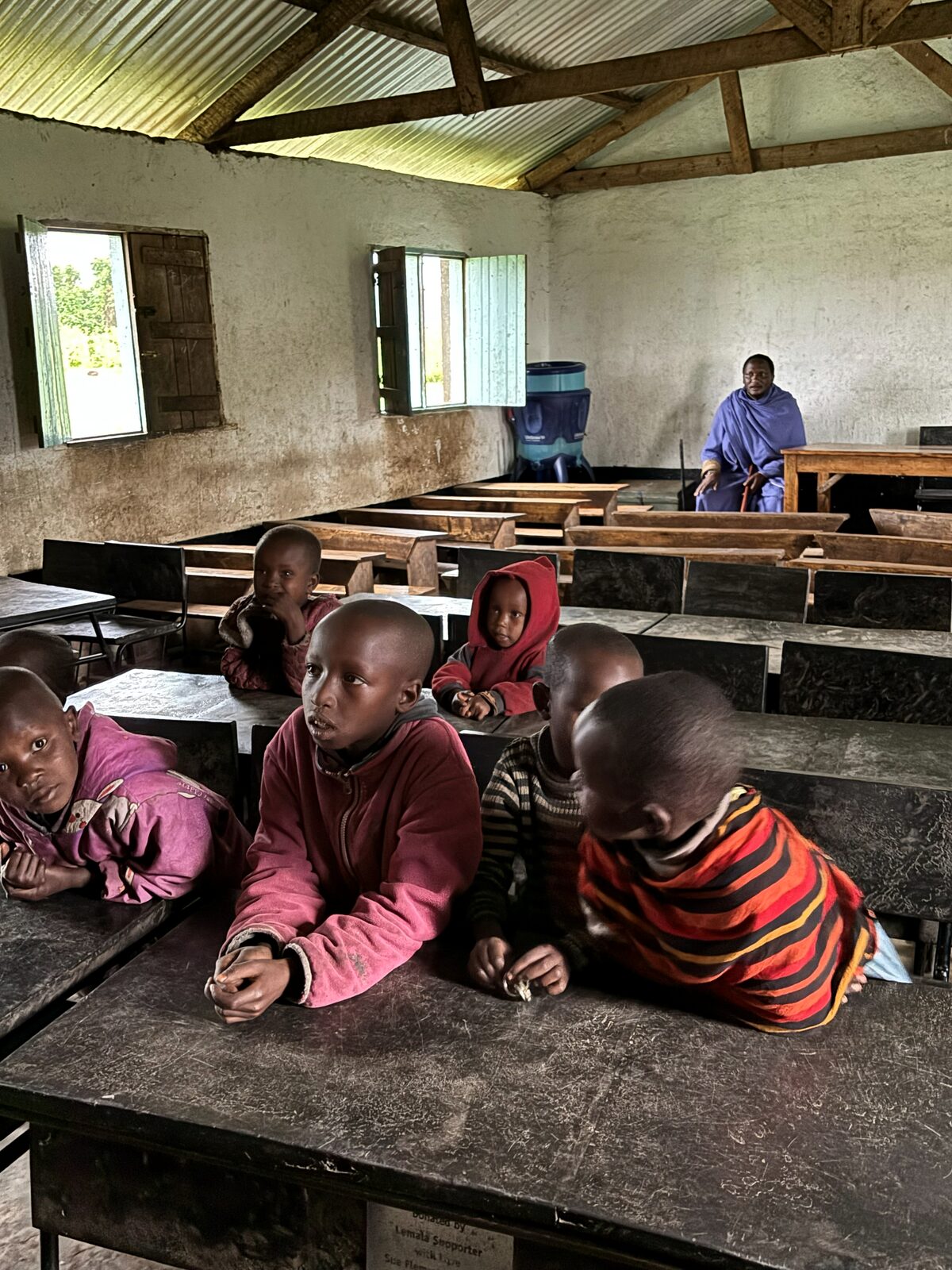 Wide view of the classroom interior, children at desks, elder seated at the front