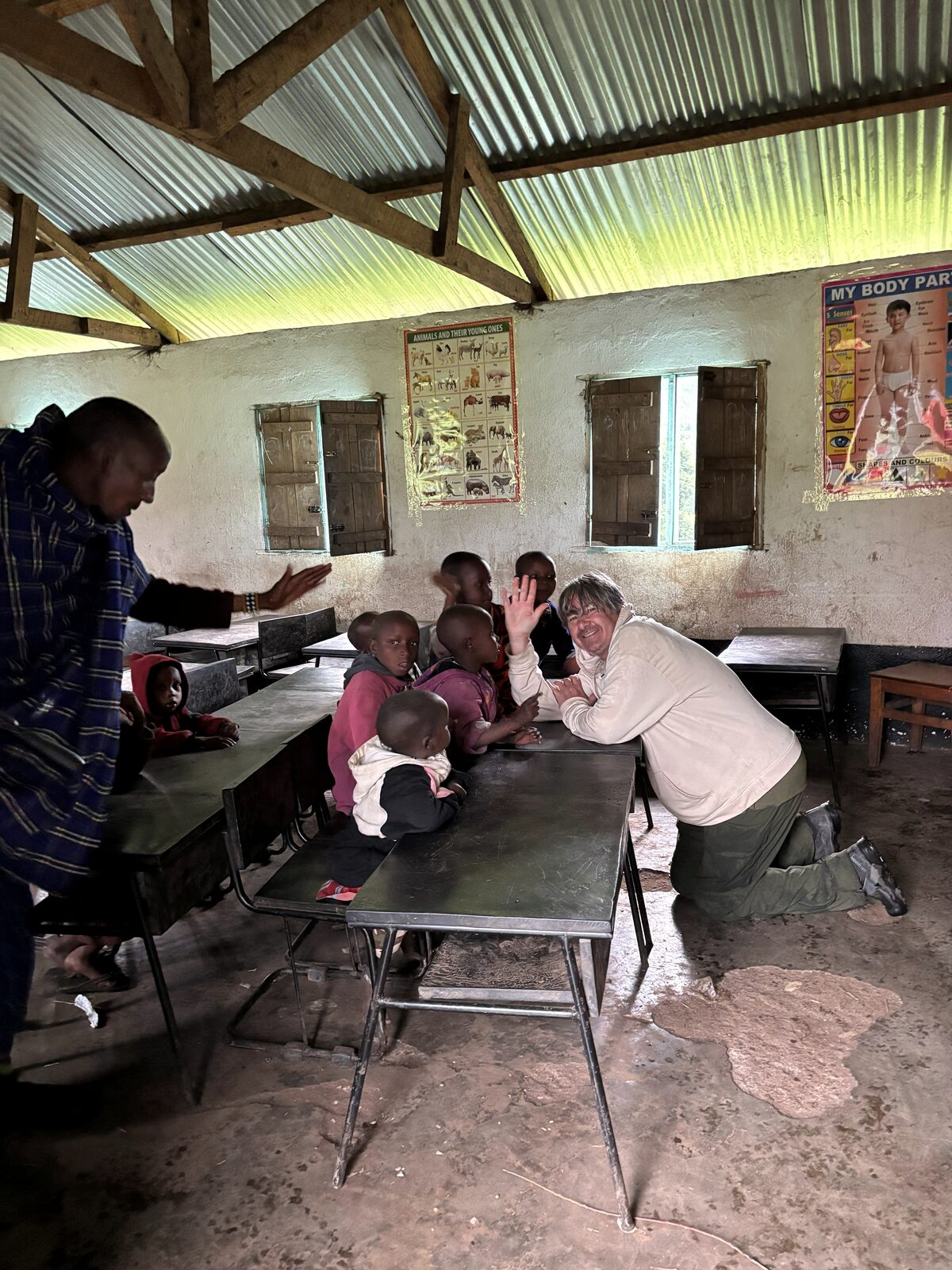 Robert kneeling at a classroom desk in a Maasai Boma, waving alongside children, Ngorongoro Conservation Area