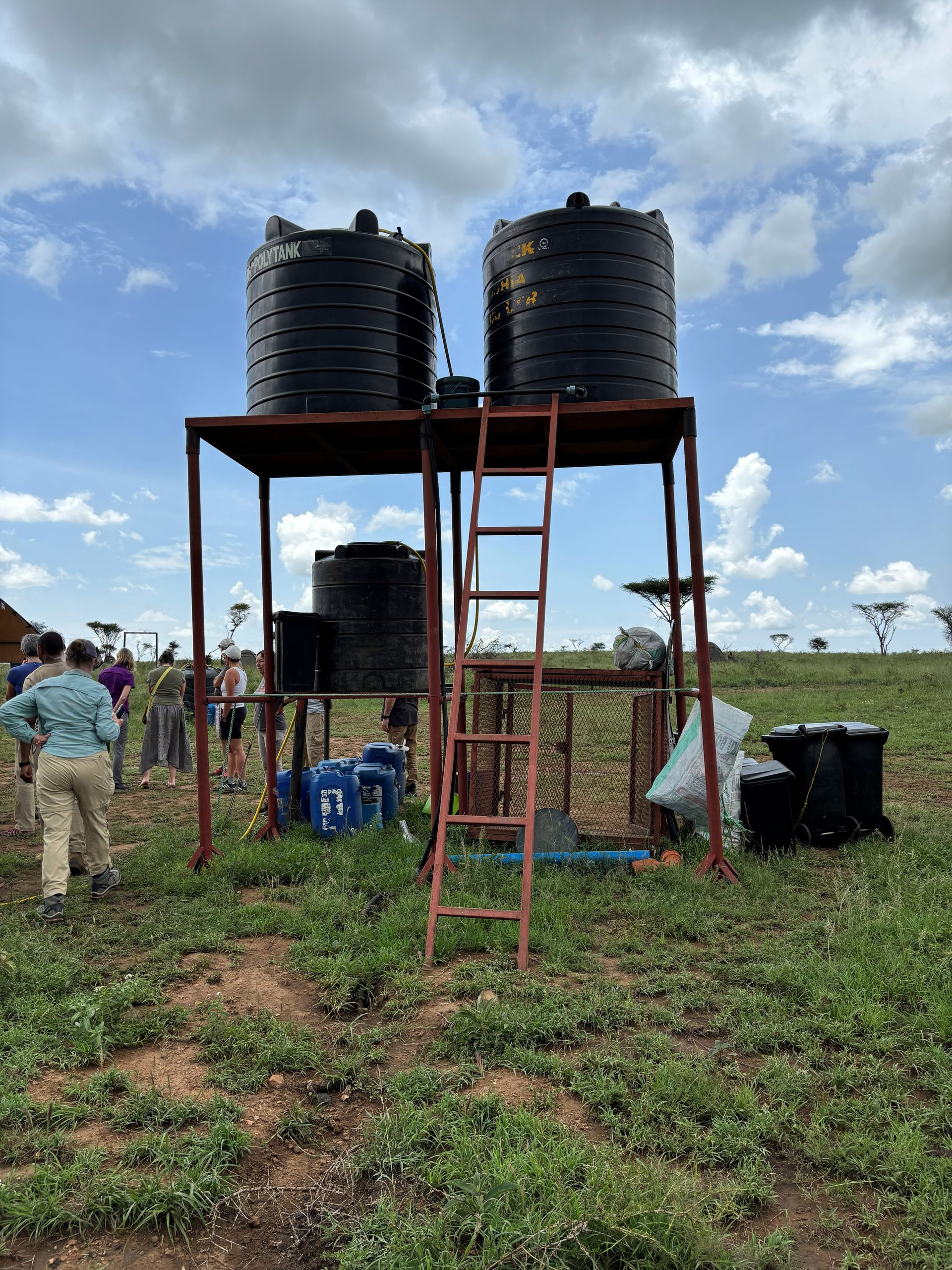 Two black polytanks on a red steel scaffold on the Serengeti plain — the camp's water supply system, with the group visible in the background