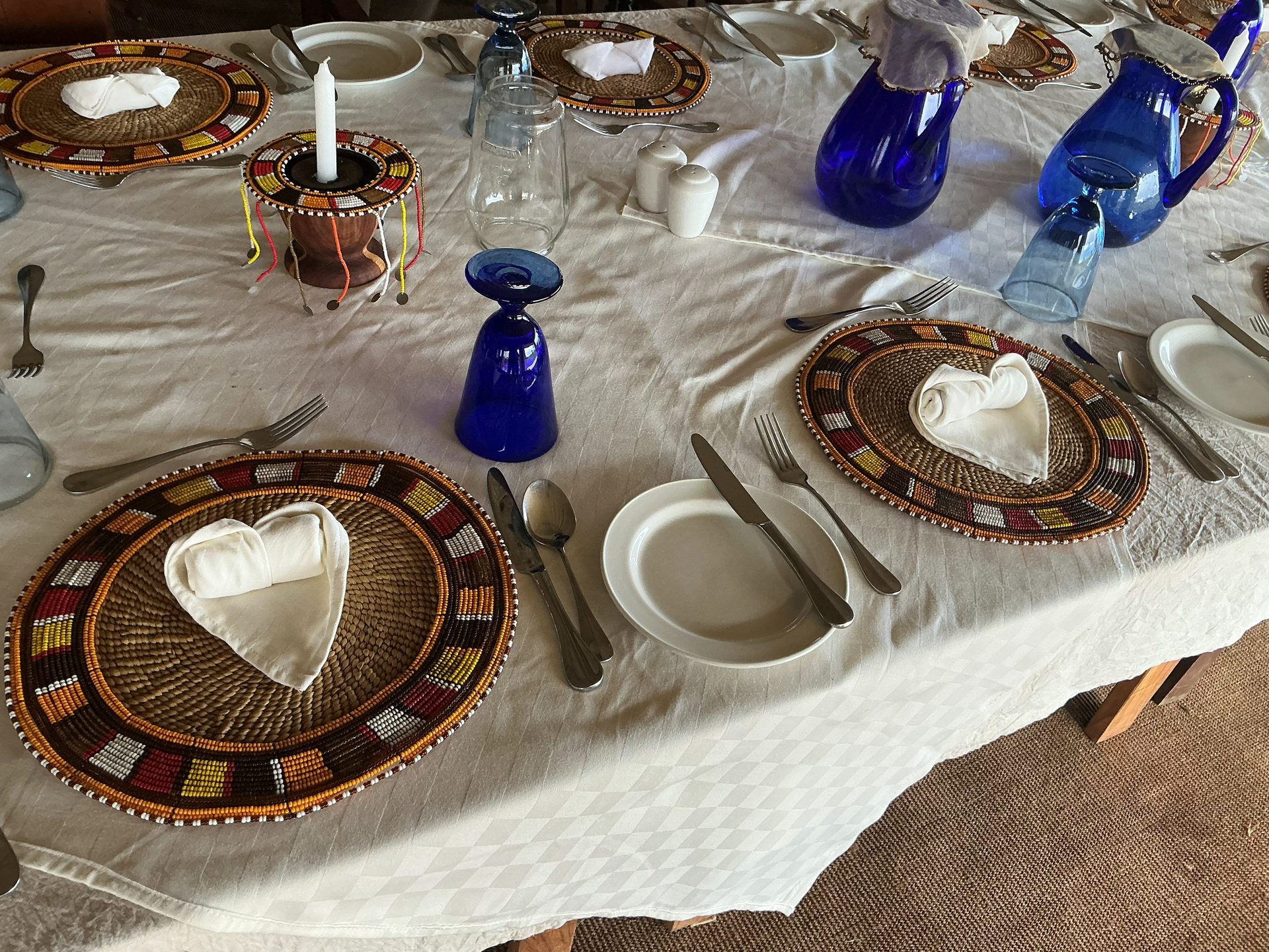 Nyumba camp dinner table set with Maasai beaded woven placemats, heart-folded white napkins, cobalt blue glassware and pitchers, and a beaded candle centerpiece