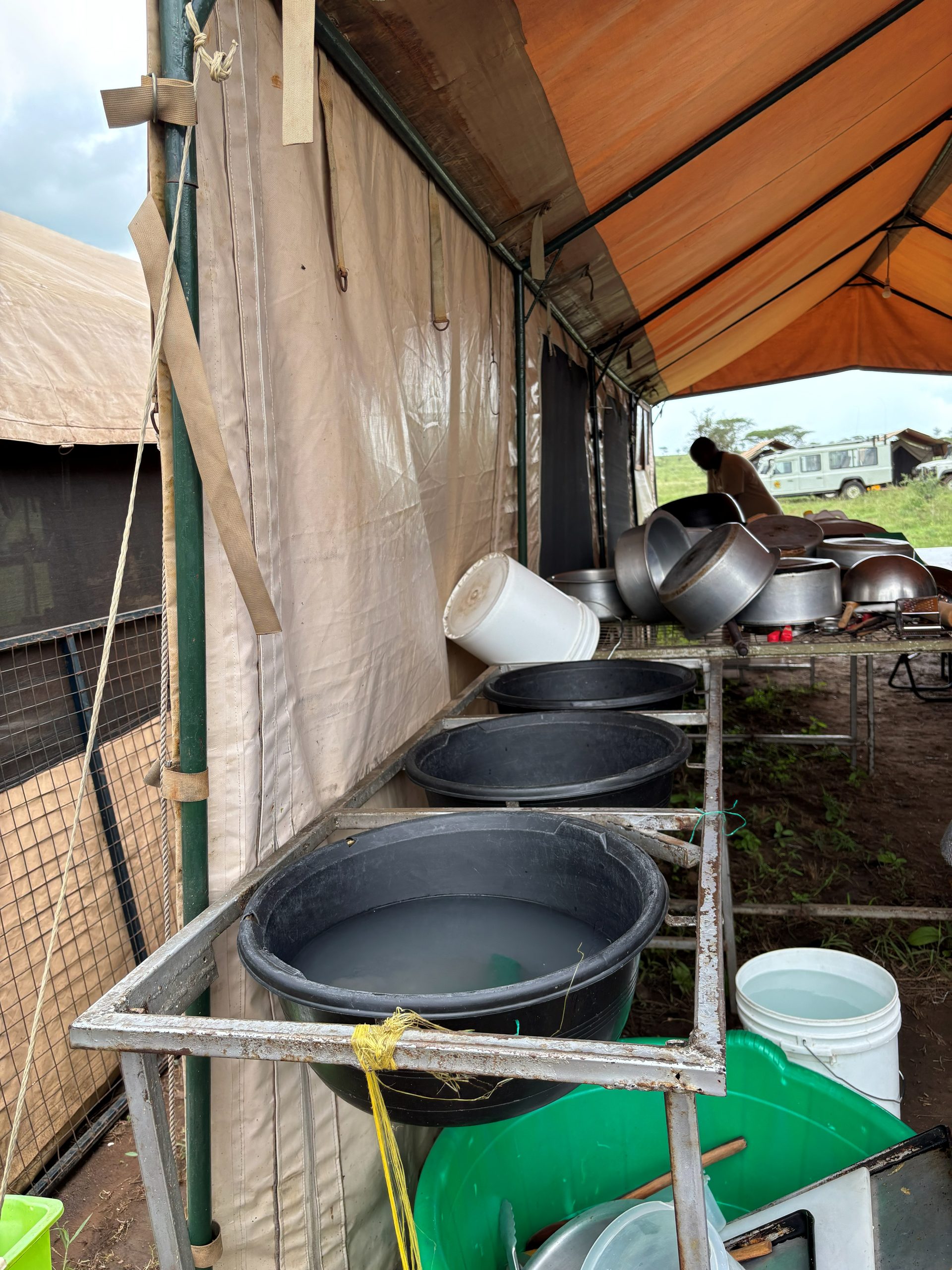 The camp dishwashing station — three large black tubs of water on a rack under the orange kitchen awning, with stacked pots behind