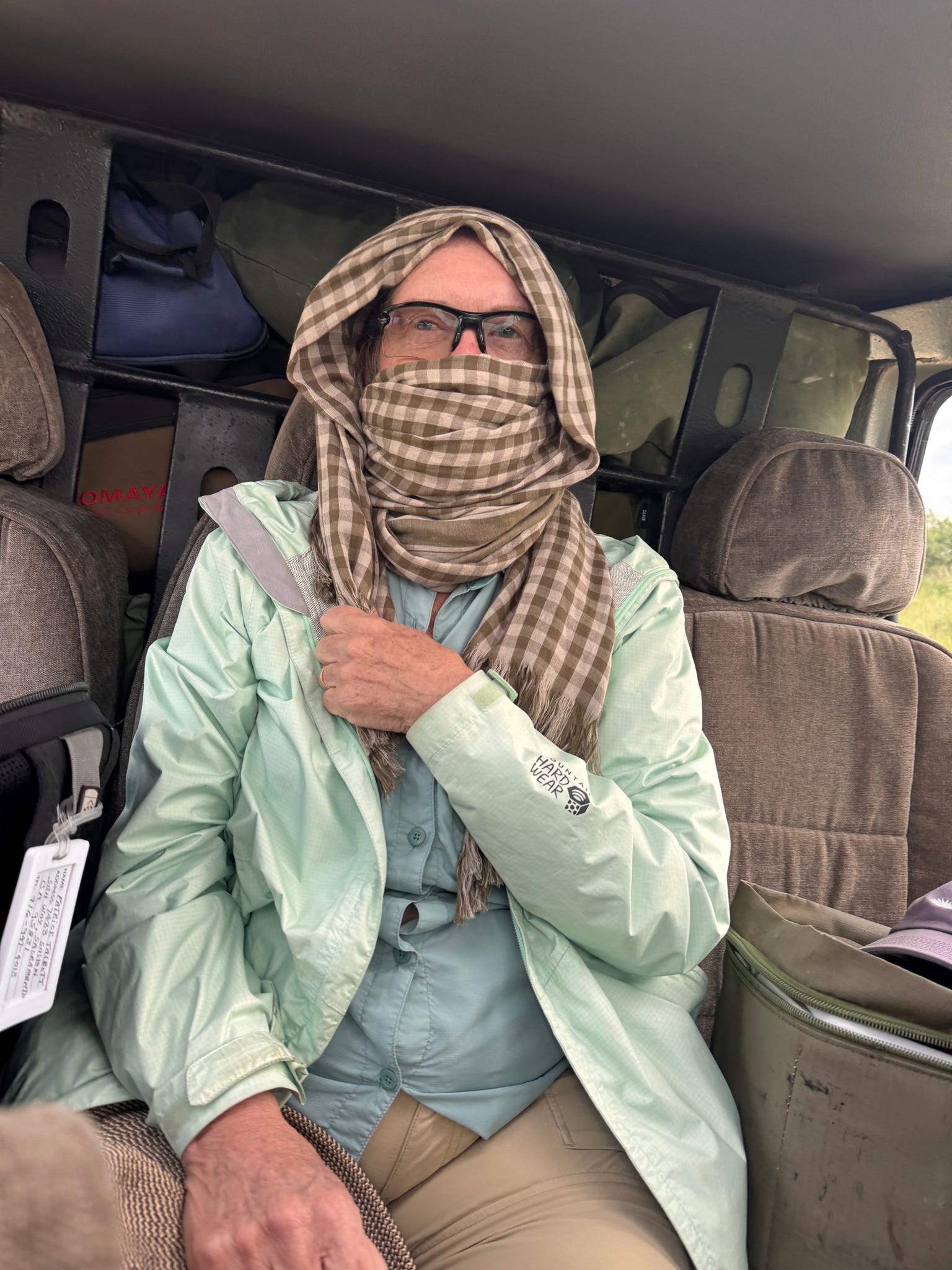 A group member in the Land Cruiser with a scarf wrapped completely around her head and face against the dust — only sunglasses visible, expression clearly resigned