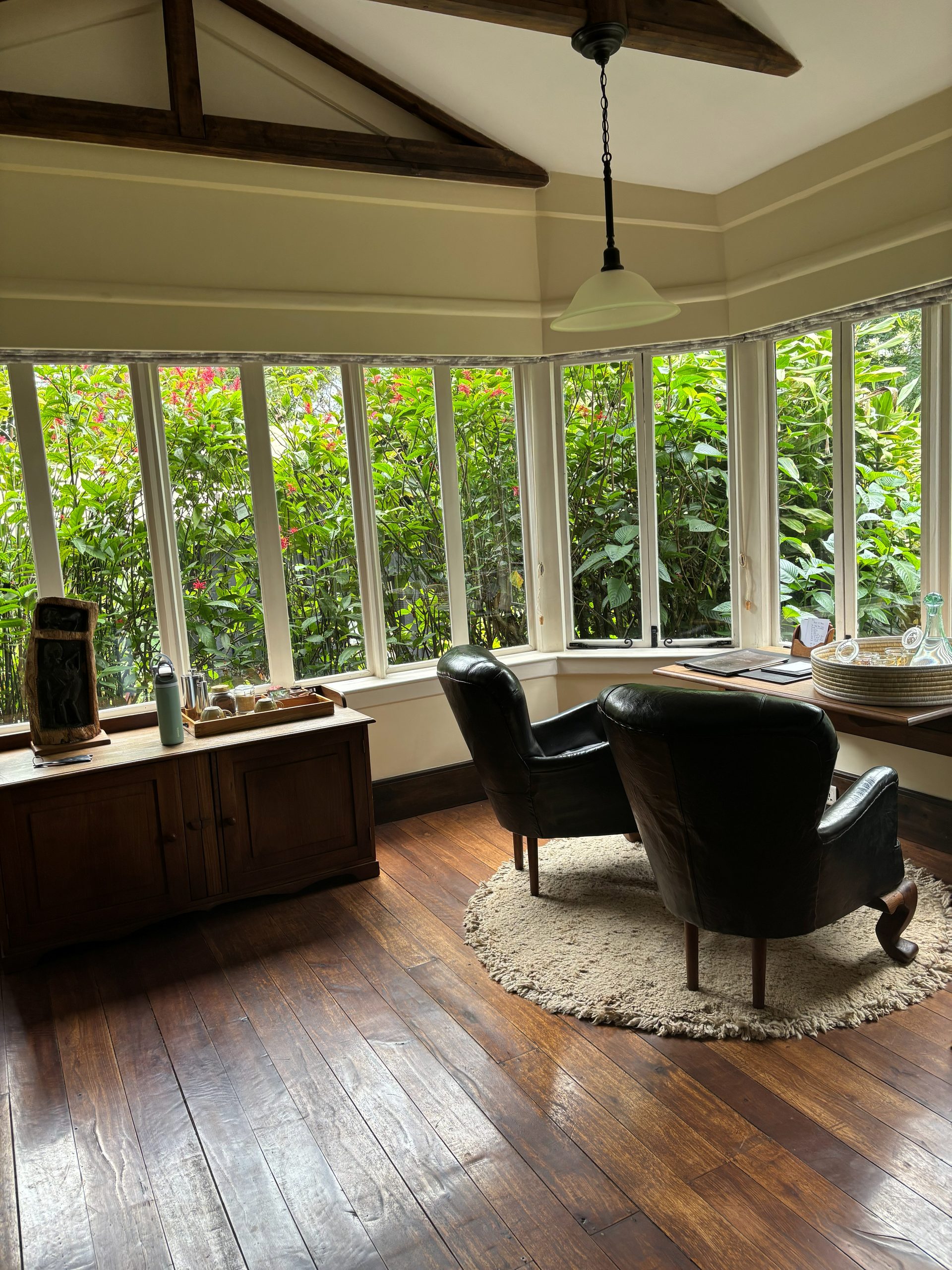 The Gibb's Farm main house sitting room — two dark leather armchairs on a round sisal rug, dark hardwood floors, a curved bow of tall white-framed windows looking directly into lush green tropical garden, timber beams overhead, pendant light, wooden sideboard with carved figure and thermos