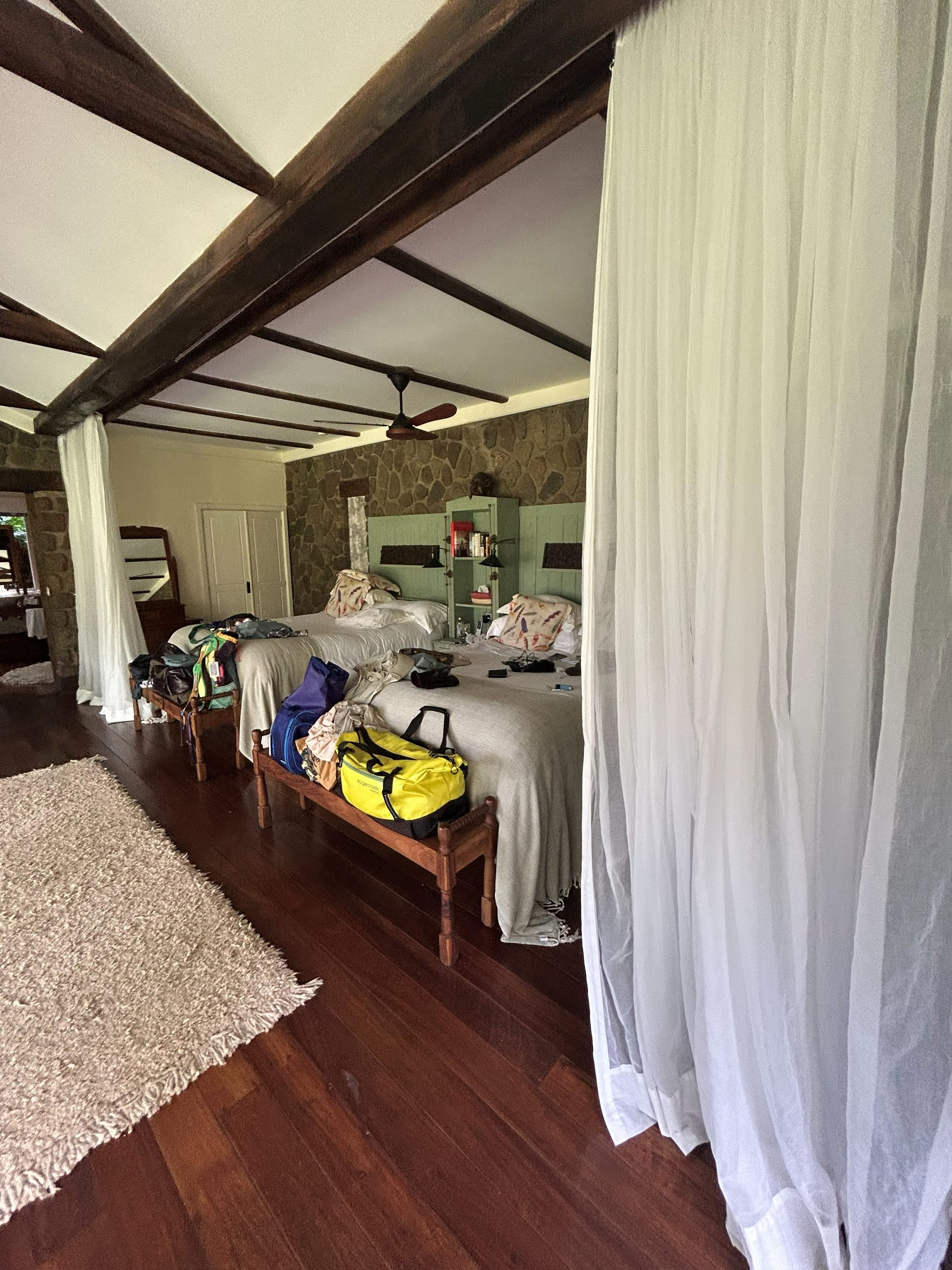 The Gibb's Farm Tloma room from the doorway — mosquito net curtains hanging from the beams, two beds with feather-print linens, dark hardwood floors, stone walls, luggage everywhere