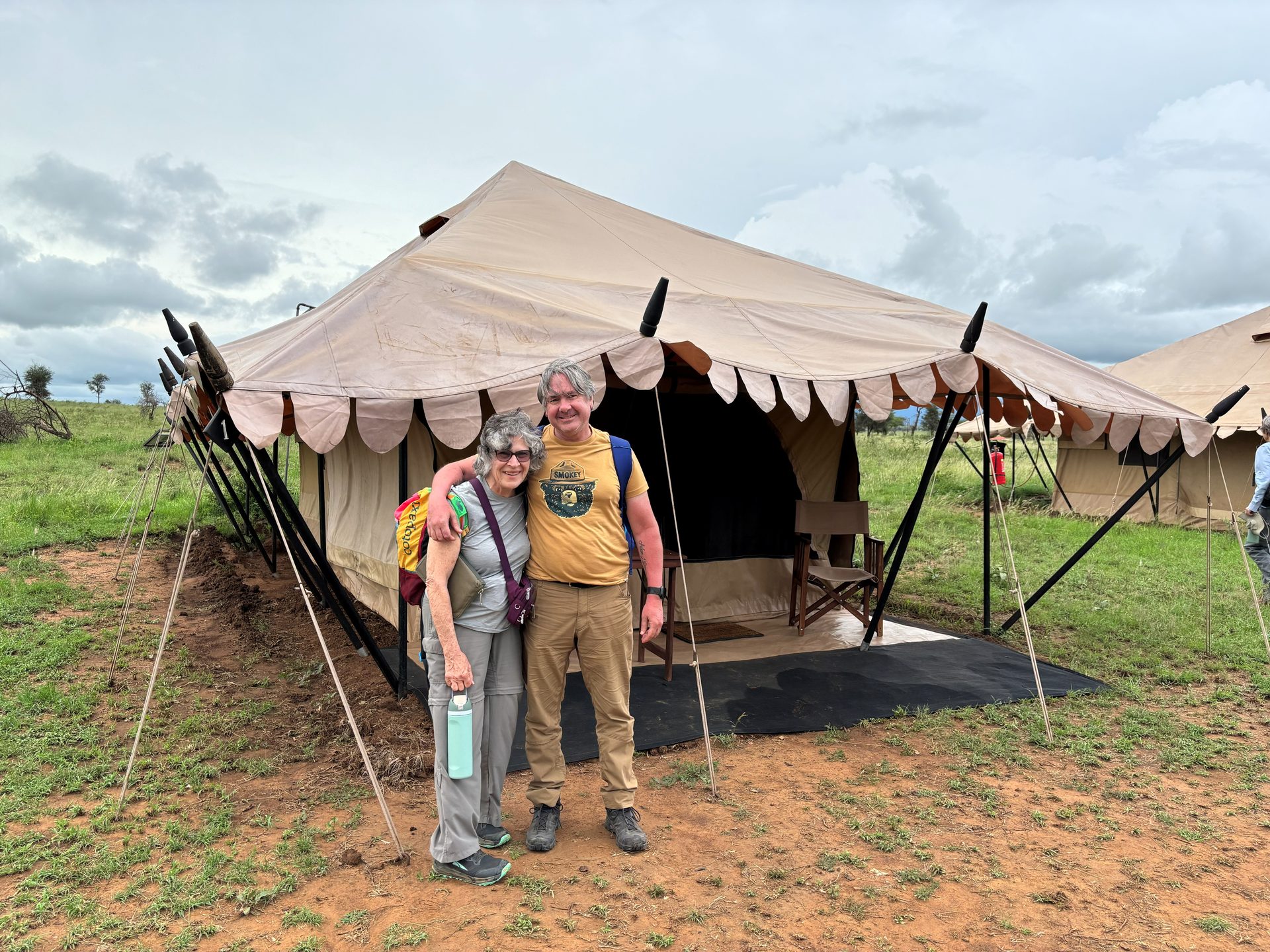 Two group members posing in front of their shared Nyumba tent — smiling, the cloudy Serengeti sky behind them
