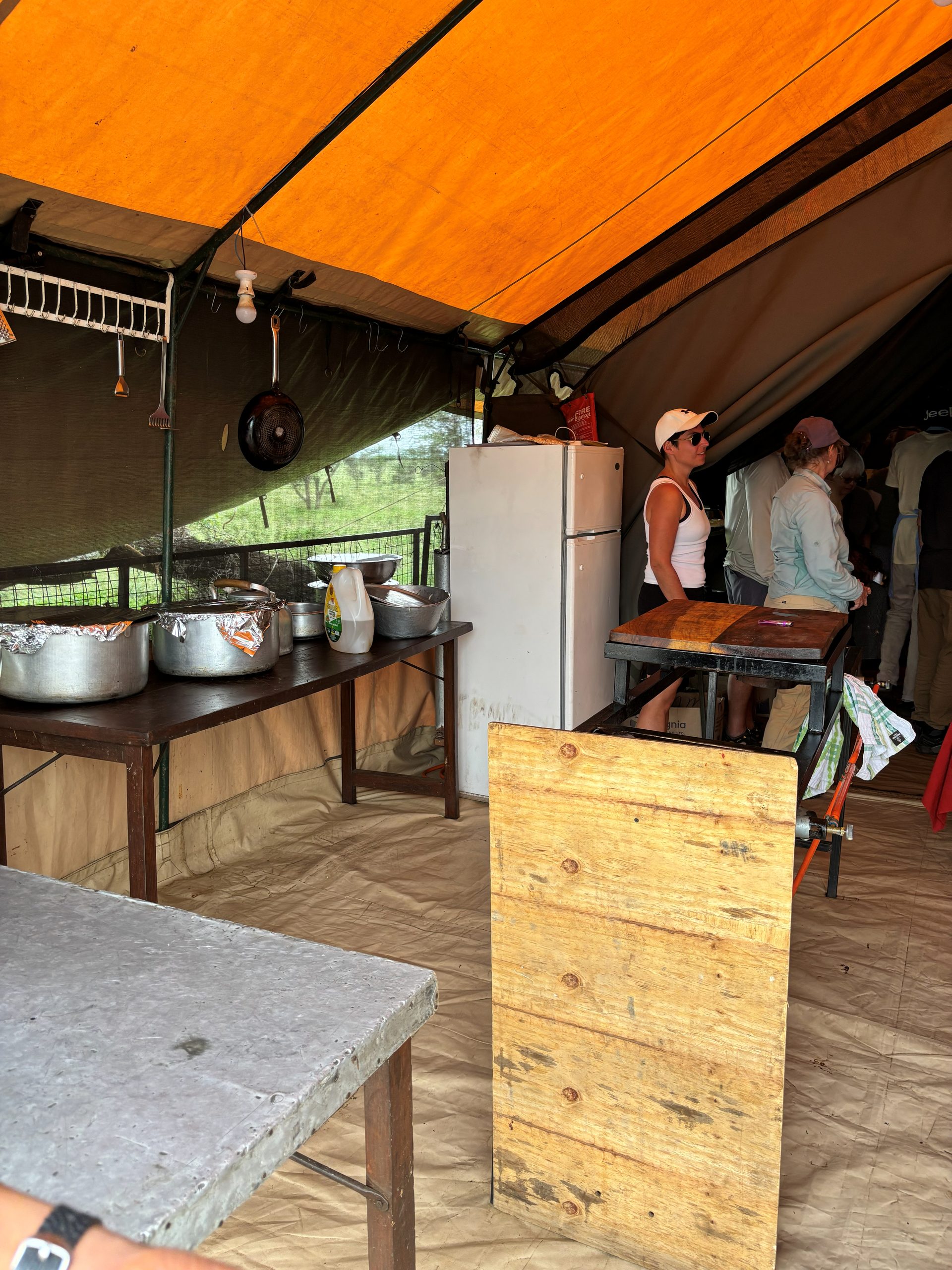 The Nyumba camp kitchen tent interior — a Thomson Safaris cook in cap stands at the prep table, a portable refrigerator behind, foil-wrapped pots lined up, tools hanging overhead