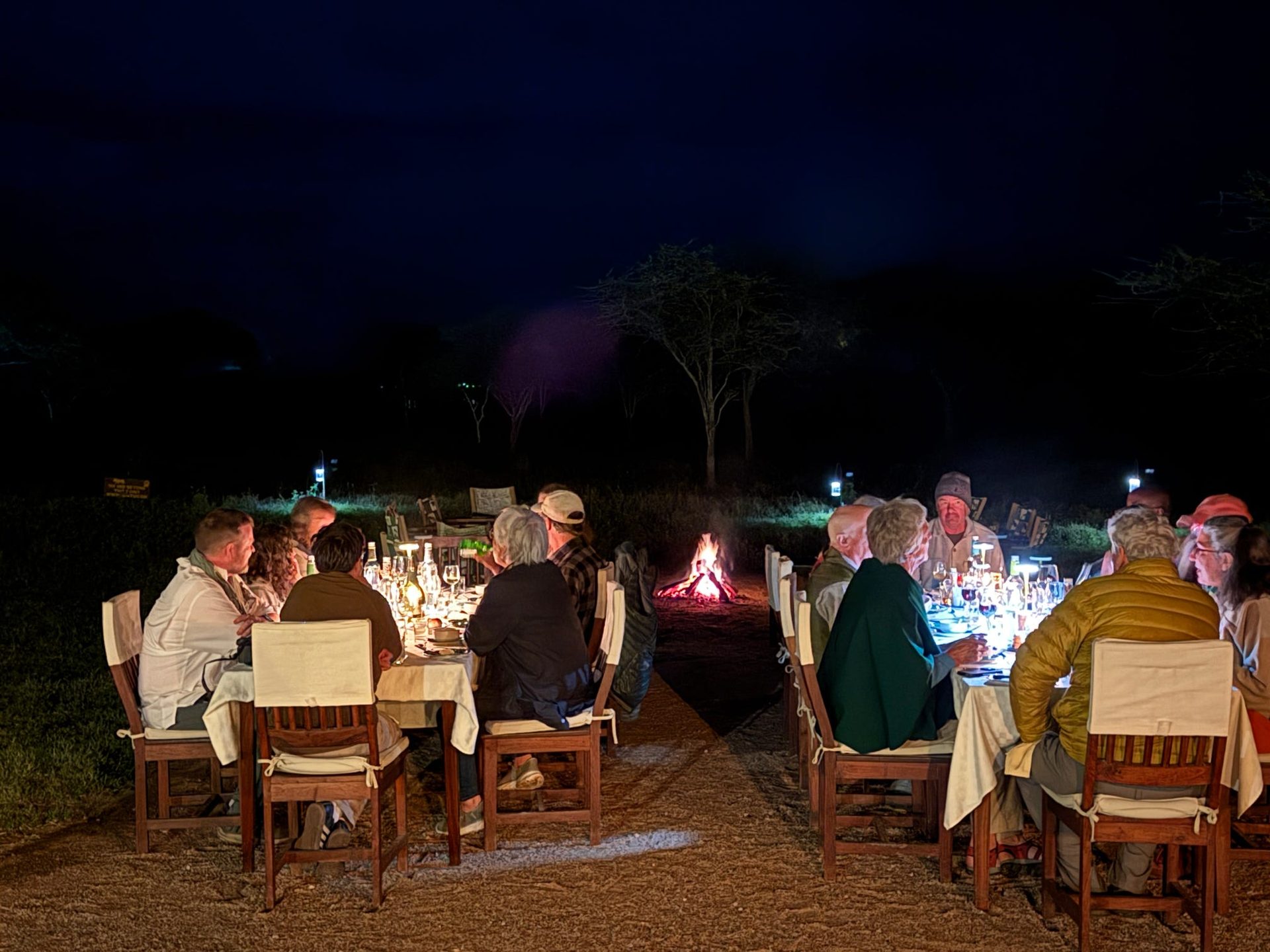 The Ndutu last night dinner table set outdoors at dusk — white linen, candlelit lanterns, napkin fans, one group member smiling wrapped in a Maasai blanket, the dark African bush behind