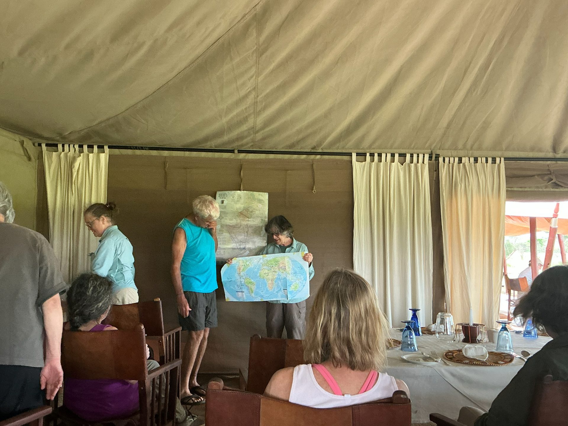 Two group members holding up a world map in the Nyumba dining tent — the group seated around, another map pinned to the canvas wall behind