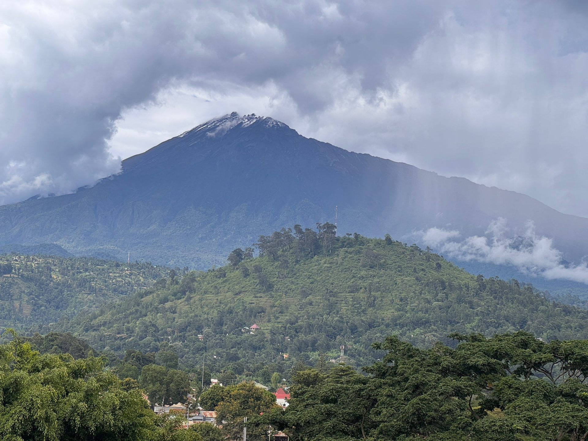 Mount Meru rising above Arusha with a rare dusting of snow on the summit — heavy clouds, green foothills, town visible below