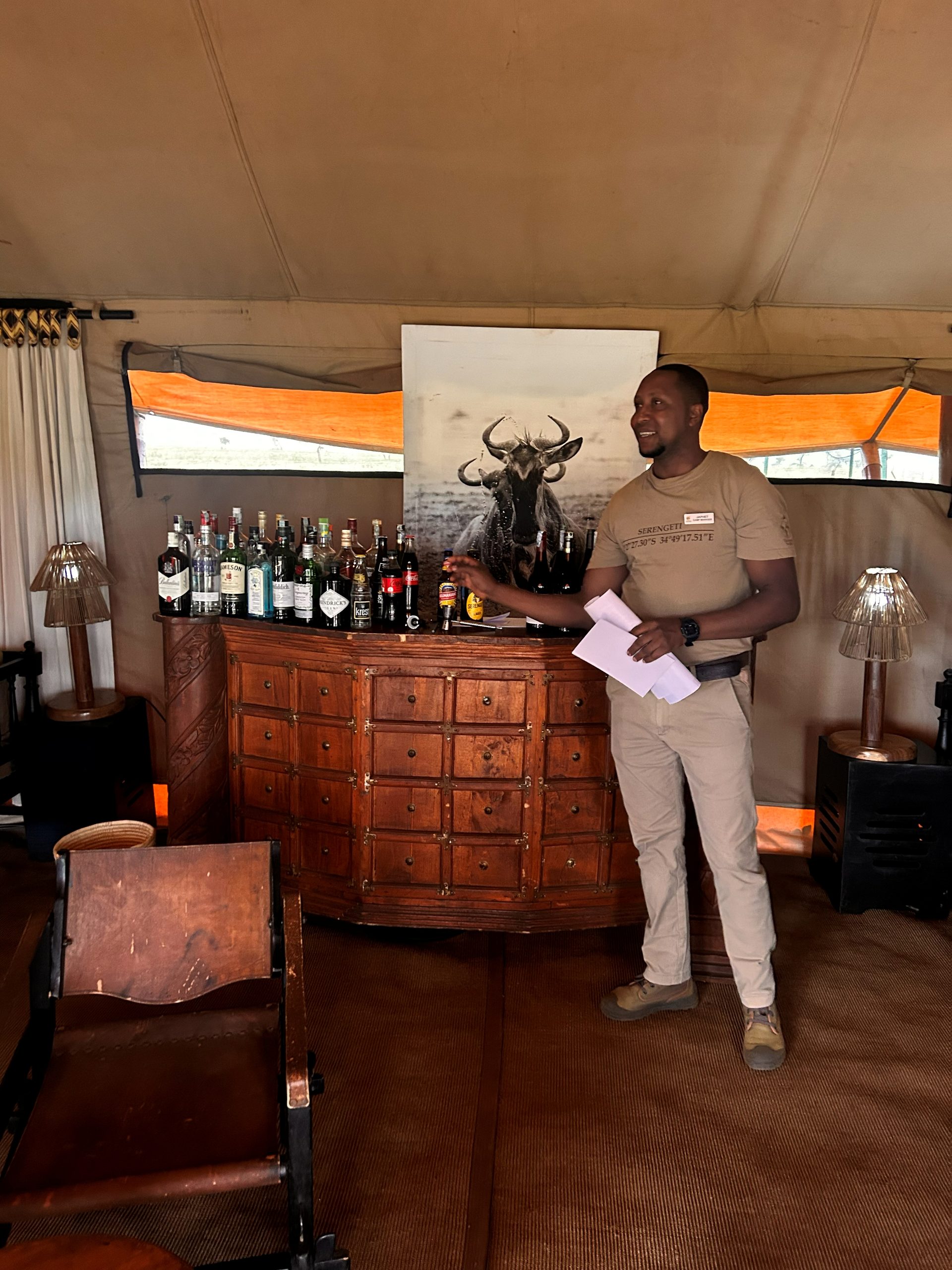 Camp manager at the Nyumba bar — carved wooden dresser stocked with bottles, wildebeest art behind