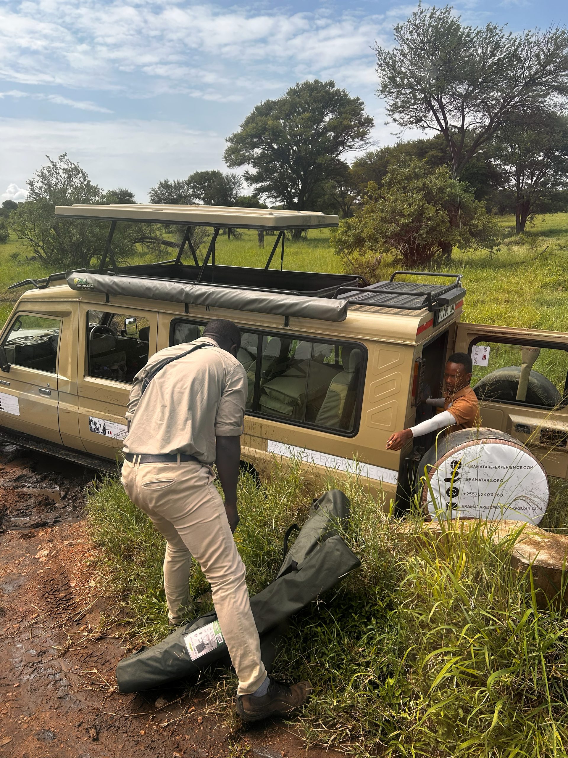 A Thomson Safaris guide pushing and steadying a Land Cruiser that has become stuck in the wet mud on a Serengeti track — another guide assists from inside