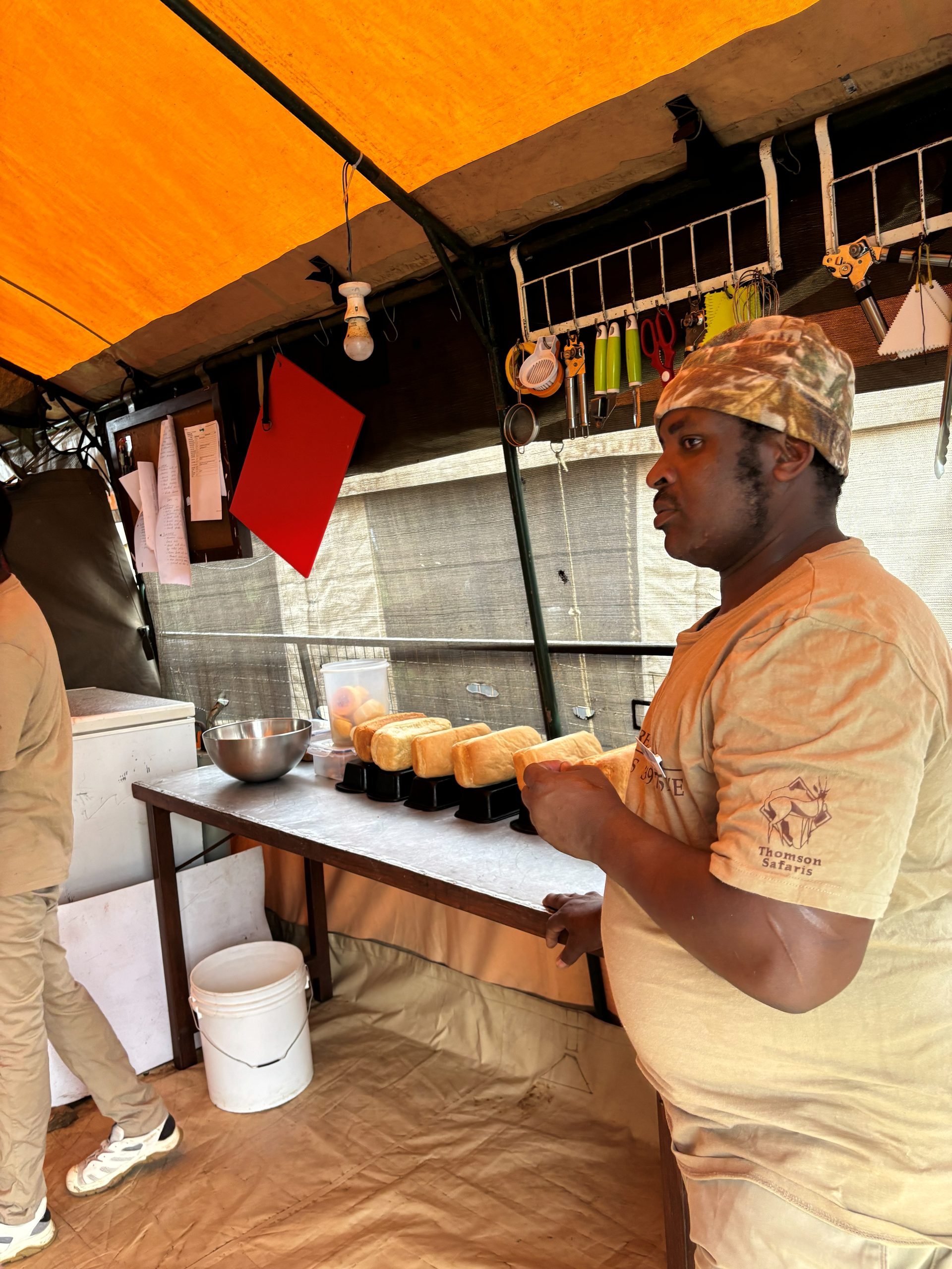 A Thomson Safaris cook in a camo bandana lining up freshly baked bread loaves on the kitchen prep table — tools and a refrigerator behind him