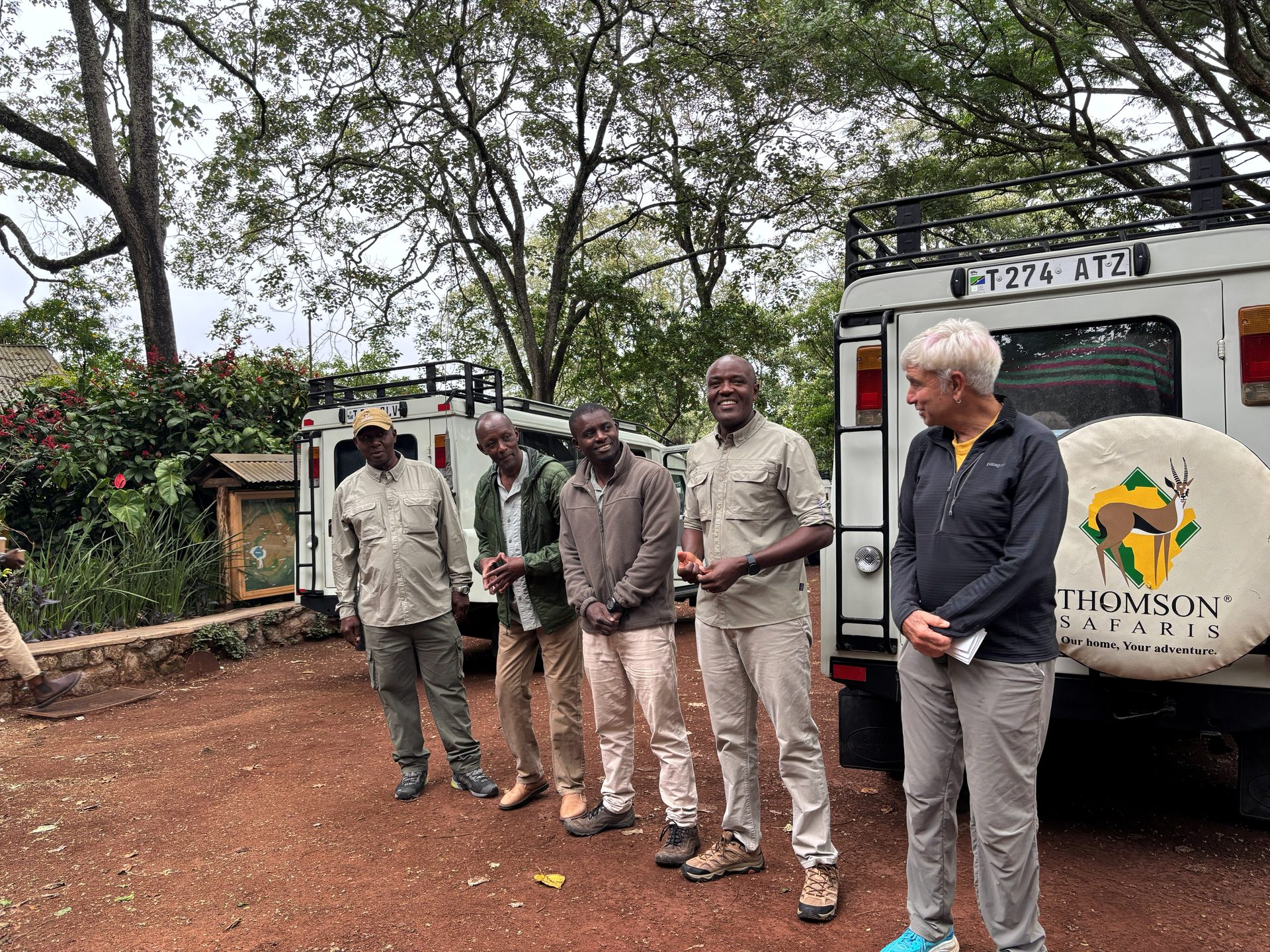 Four Tanzanian Thomson Safaris game driver-guides standing together beside the Land Rover at Gibb's Farm — all smiling broadly, one group member with white hair standing beside them, the Thomson Safaris spare tire cover and lush garden visible behind