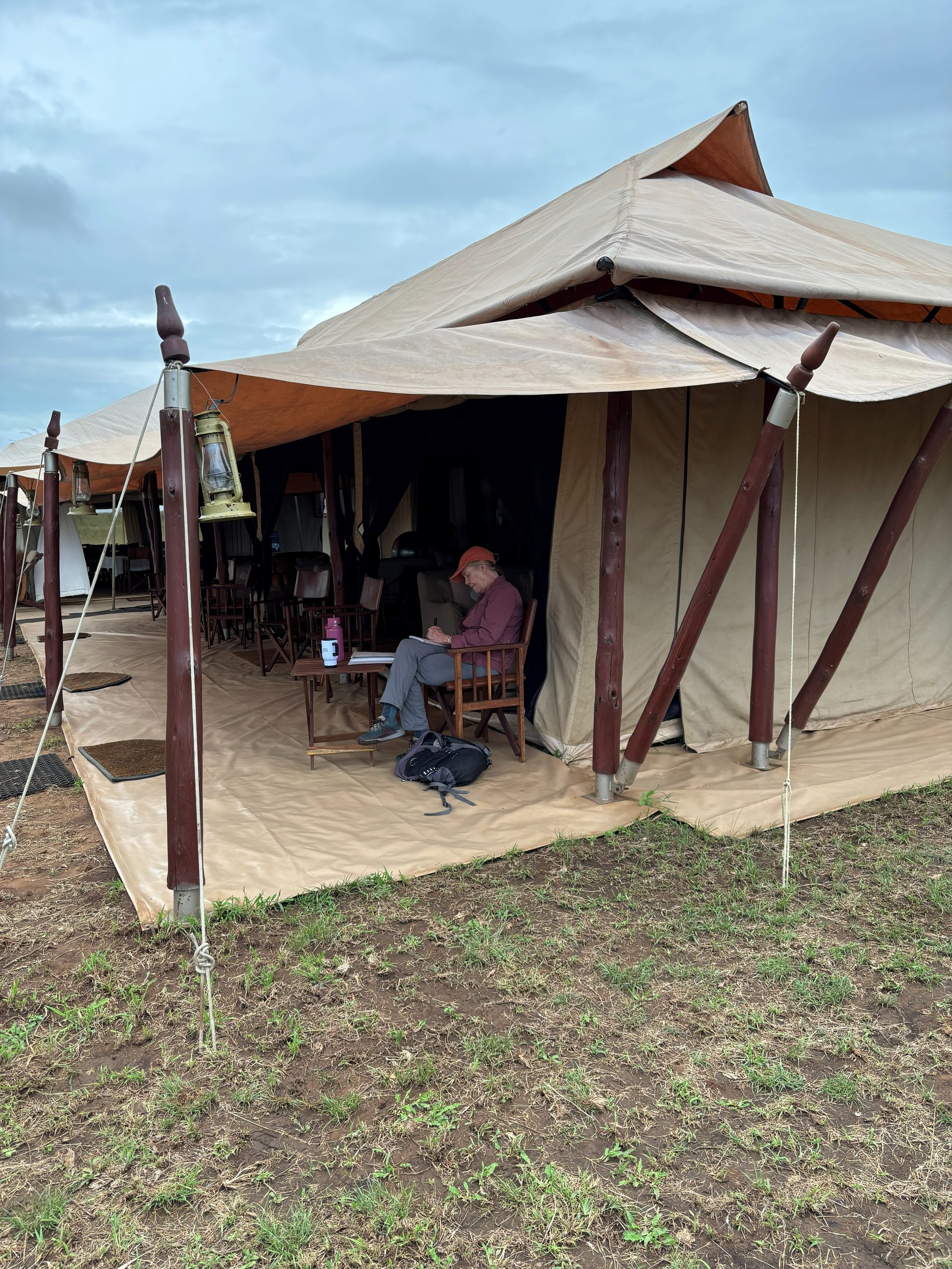 A group member sitting alone at the entrance of the dining tent, writing in a journal at a small table, a lantern hanging beside them